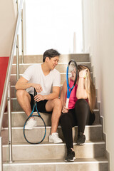 Pareja multirracial sentados conversando en las escaleras después de jugar un partido de tenis - obrazy, fototapety, plakaty
