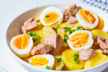 Tuna and potato salad with egg and green onions in white bowl on white tile background. Healthy vegetarian food, fitness meal.