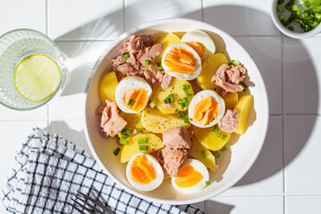 Tuna and potato salad with egg and green onions in white bowl on white tile background. Healthy...
