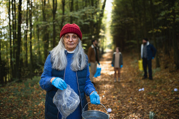 Senior woman volunteer looking at camera when cleaning up forest from waste, community service.