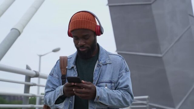 Modern devices to listen music. Stylish African-American guy in knitted hat with earphones and cellphone walks along footbridge in large city spbas