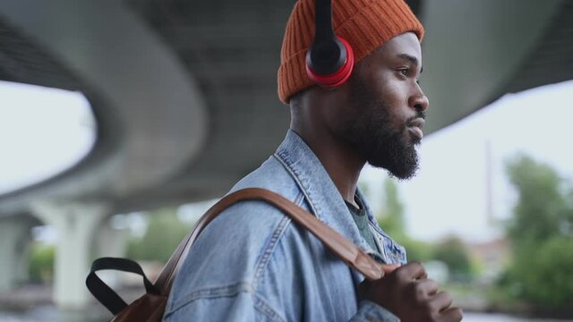 Music lover walking along street. Thoughtful african-american guy model with spbas wireless earphones and urban backpack walks under river bridge on embankment closeup