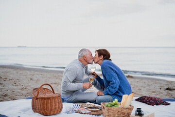 Happy senior couple in love sitting on blanket and kissing when having picnic outdoors on beach.