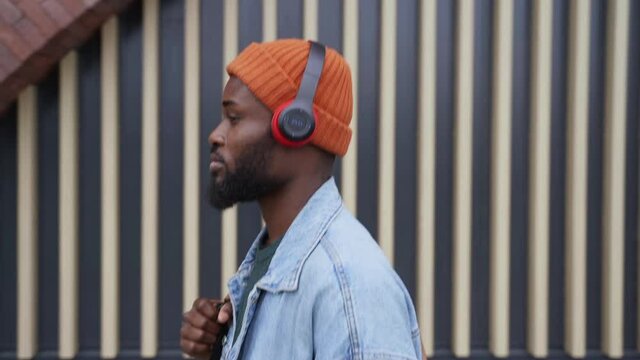 Music lover walking along street. Handsome African-American person with modern earphones walks passing large building wall in city street side view spbas