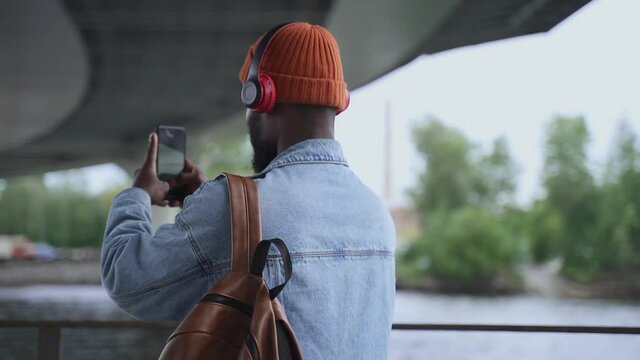 Contemporary gadgets to enjoy life. African-American man tourist with spbas wireless headphones takes picture of river by phone under bridge backside view