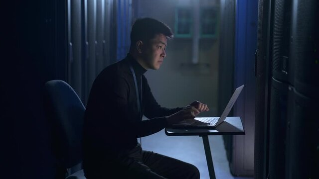 Male engineer working with laptop and inspecting equipment, sitting at table in data office spbas. Side view of young man checks hardware and looks at computer screen, writes report and sits in dark