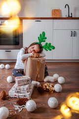 Fototapeta na wymiar Little girl with big christmas gift and lights. Gift boxes and christmas toys on the ooden floor. Kid in deer horns.