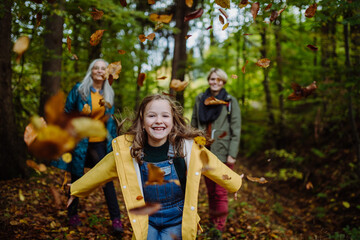 Happy little girl running and throwing leaves during autumn walk with mother and grandmother in forest