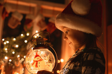 little girl looks at a Christmas lantern illuminating the face in Santa's hat at night in the room,