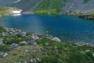 Fototapeta na wymiar Aerial drone view of alone woman traveler in dress and hat watching on Seven colored mountain glacial lake, summer landscape: high peaks, snow, stones. Arkhyz, Caucasus, Karachay-Cherkessia, Russia