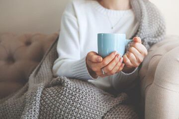 Close up of hands holding blue cup of tea or coffee. Model in white sweater and cozy plaid is sitting, relax at home on sofa. Depth of field and empty space on blurry beige background