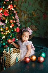 Fototapeta na wymiar a little African-American girl in a pink dress with Christmas balls near the Christmas tree. the child decorates