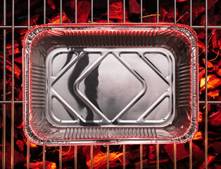 Empty aluminum foil food container on bbq grate over hot pieces of coals. Top view.