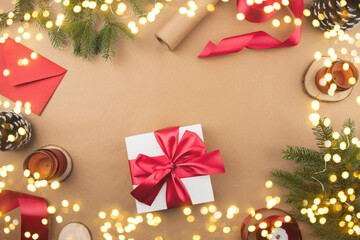 White gift box with red ribbon on a kraft background. Packaging and preparation of gifts for the celebration. Christmas New Year backdrop with tree branch decor and lights