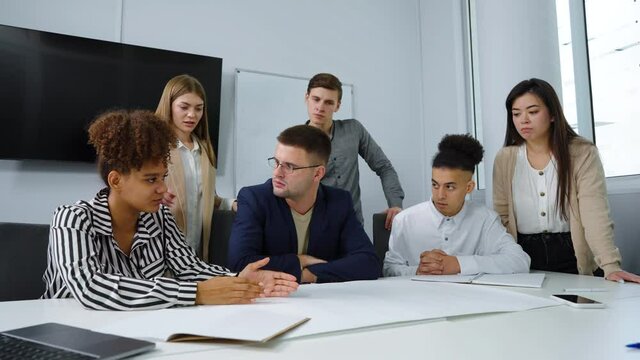 Group of designers gathering in office and discussing masterplan lying on table, boss sitting in center and listening to ideas of employees. Tracking shot multiethnic group brainstorming