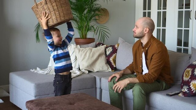 Father and toddler on laundry day. Little son carries large wicker basket of spbd washed clothes to unload for parent sitting on sofa in living room