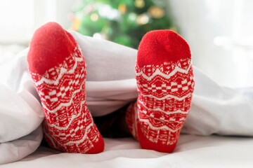Cute kids feet in red knitted woolen socks covered with blanket on white bed near Christmas tree. Cozy Merry Christmas morning at home, happy moments of winter holidays, Christmastime concept.