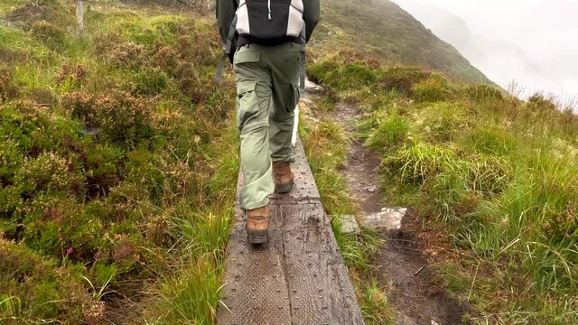 Slow motion rear view of hiker with dog walking on narrow wooden trail in high greened mountains of Ireland - Rainy day with dense fog in the valley