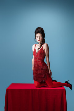 Portrait of young adorable woman in dark red, wine color evening dress posing on table with red cloth isolated on blue background.