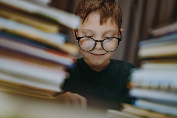 cute caucasian boy wearing glasses reading book. Cheerful boy peeking from behind piles of books