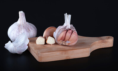 A head of garlic, a few cloves of young fresh garlic in a transparent husk, a chopped clove of garlic on a wooden cutting board. Black background