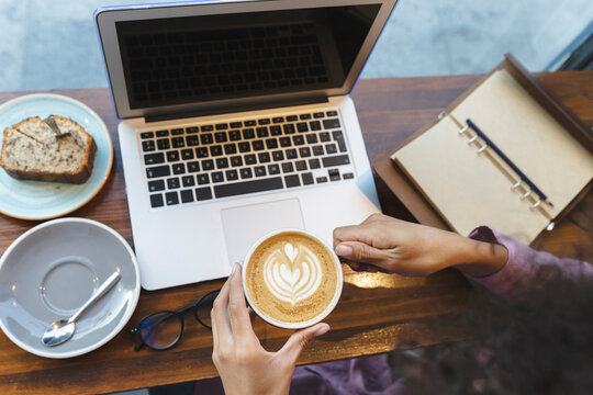 Businesswoman with laptop holding cappuccino cup at cafe table