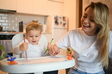 Little cute baby toddler boy blonde sitting on baby chair learning to draw. Beautiful young mom and son playing spend time together indoors at home with toys. Healthy happy family childhood concept