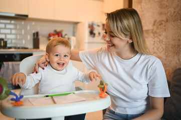 Little cute baby toddler boy blonde sitting on baby chair learning to draw. Beautiful young mom and son playing spend time together indoors at home with toys. Healthy happy family childhood concept