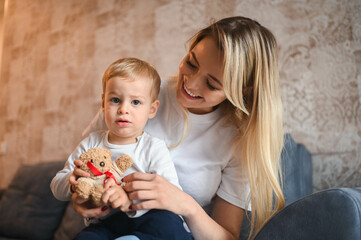 Little cute baby toddler boy blonde sitting on mother's arms. Beautiful young mom and son playing spend time together indoors at home with toys. Healthy happy family childhood concept