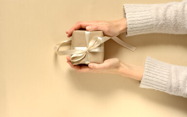 Female hands in a sweater giving a gift wrapped in natural paper and a satin ribbon on a beige background.