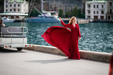 A woman in a red dress, a fashion model with long silk wings in a flowing dress, flying fabric on the embankment.