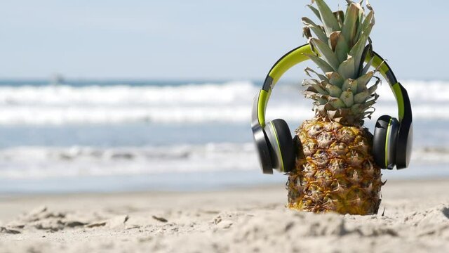 Funny pineapple in headphones, sandy ocean beach, blue sea water waves, California pacific coast, USA. Tropical summer exotic fruit enjoying vacations and music in paradise. Ananas sunbathing on shore