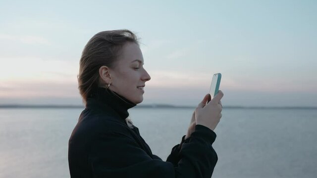 A woman takes pictures on her phone of a lake at sunset on an autumn evening. Taking video of a sunset over the lake. Woman loves nature. High quality 4k footage