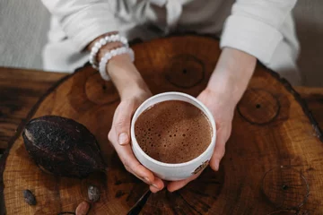Foto op Canvas Hot handmade ceremonial cacao in white cup. Woman hands holding craft cocoa, top view on wooden table. Organic healthy chocolate drink prepared from beans, no sugar. Giving cup on ceremony, cozy cafe © ninelutsk