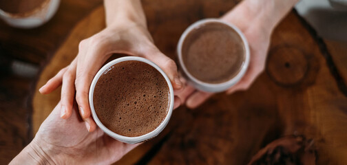 Hot handmade ceremonial cacao in white cups. Woman hands giving craft cocoa, top view on wooden table. Organic healthy chocolate drink prepared from beans, foam Giving cup on ceremony cozy atmosphere
