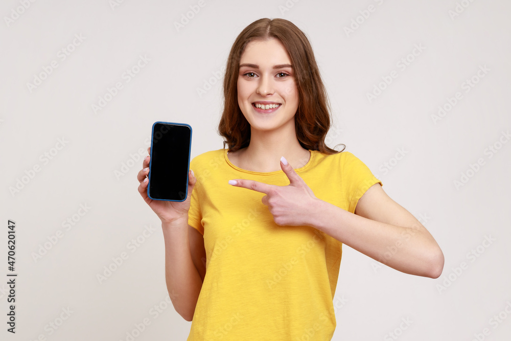 Wall mural Look at my cellphone! Adorable glad teenager girl pointing smartphone with finger and smiling at camera, advertising of mobile device. Indoor studio shot isolated on gray background.