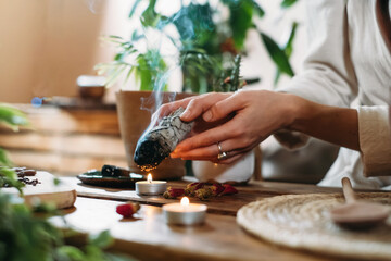 Woman hands burning white sage, palo santo before ritual on the table with candles and green plants. Smoke of smudging treats pain and stress, clear negative energy