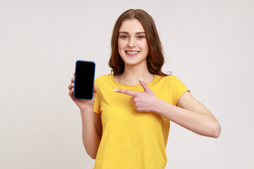 Look at my cellphone! Adorable glad teenager girl pointing smartphone with finger and smiling at camera, advertising of mobile device. Indoor studio shot isolated on gray background.