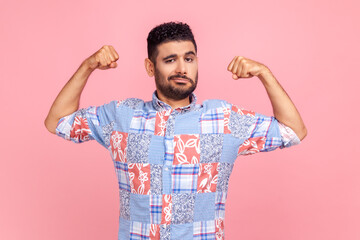 I'm strong! Portrait of attractive man in casual blue shirt raising hands to show biceps and looking confident at camera, feeling power and strength. Indoor studio shot isolated on pink background.