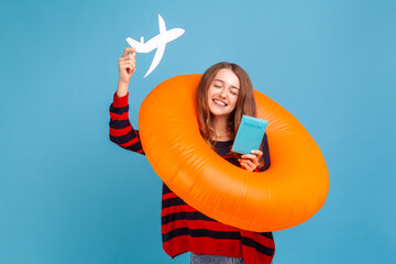 Extremely happy woman wearing striped casual style sweater, standing with rubber ring, holding passport and airplane mockup, rejoicing travel tour. Indoor studio shot isolated on blue background.