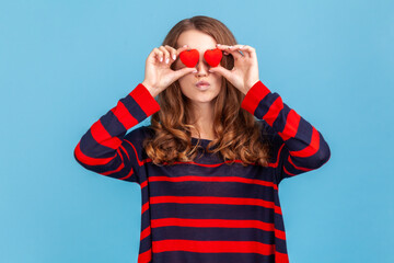Romantic woman wearing striped casual style sweater, standing with pout lips, sending air kisses, covering eyes with small red heart love symbols. Indoor studio shot isolated on blue background.