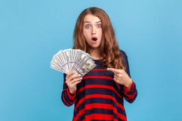 Shocked woman wearing striped casual style sweater, pointing at dollars banknotes in her hand, having astonished expression, big profit. Indoor studio shot isolated on blue background.