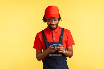 Smiling worker wearing uniform reading message on mobile phone in online delivery order app, house repair maintenance or delivery services. Indoor studio shot isolated on yellow background.