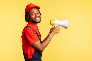 Side view portrait of smiling happy handyman wearing blue overalls and red cap, holding megaphone, looking at camera with toothy smile. Indoor studio shot isolated on yellow background.
