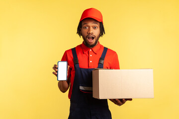 Portrait of surprised astonished courier wearing red T-shirt and blue overalls holding cardboard box and cell phone with empty screen. Indoor studio shot isolated on yellow background.