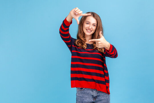 Smiling woman wearing striped casual style sweater looking at camera through hand frame, capturing photo or focusing eyesight on target. Indoor studio shot isolated on blue background.