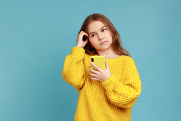 Portrait of little girl holding smart phone in hands, looking away with thoughtful facial expression, wearing yellow casual style sweater. Indoor studio shot isolated on blue background.