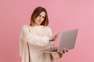 Naklejka premium Woman showing stop gesture to laptop screen, warning with prohibition sign when talking on video call, online communication, wearing white sweater. Indoor studio shot isolated on pink background.