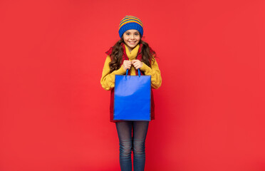 cheerful kid in winter hat hold shopping bags on red background, christmas shopping
