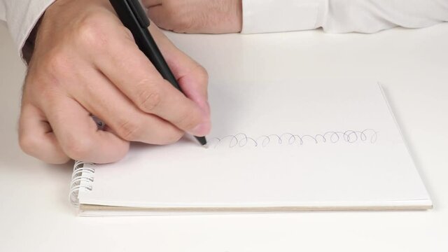 The hands of a young man in a white shirt are drawing squiggles in a white notebook